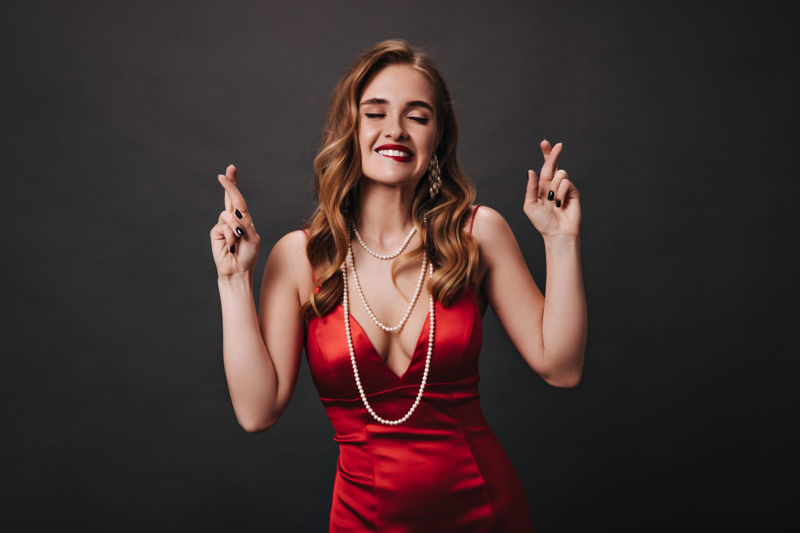 Cool lady in red silk outfit crosses fingers on black background. Blonde curly woman in satin dress